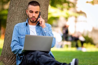 Genç adam dışarıda çalışmak ya da çalışmak için dizüstü bilgisayar kullanıyor. Genç bir adam günlük giysiler giyip halka açık bir parkta çimlere oturuyor ve bir telefon görüşmesi yapıyor..
