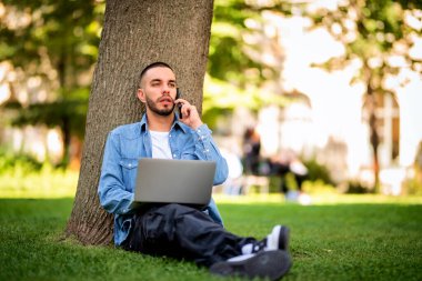 Genç adam dışarıda çalışmak ya da çalışmak için dizüstü bilgisayar kullanıyor. Genç bir adam günlük giysiler giyip halka açık bir parkta çimlere oturuyor ve telefonla konuşuyor. Tam boy çekim.