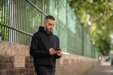 Şehirde sokakta dikilirken yakışıklı bir gencin cep telefonunu kullanırken çekilmiş bir fotoğrafı. Kapüşonlu ve kısa mesajlı kendinden emin bir erkek.. 
