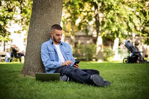 屋外で勉強したり働いたりするためにラップトップを使用する若者 カジュアルな服を着て 公園やテキストメッセージで草の上に座っている若い男性 全長ショット ストック写真