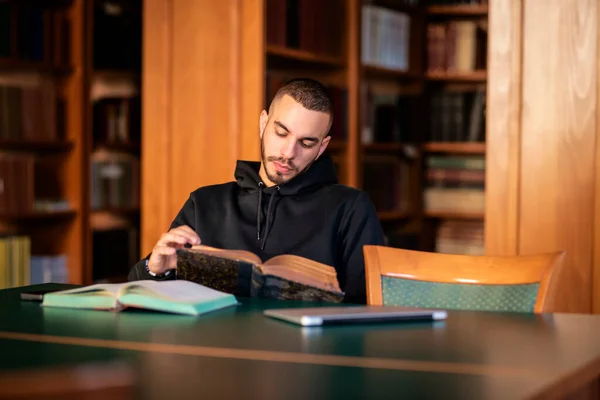 坐在大学图书馆学习的年轻人 英俊的男人穿着帽衫看书 图库图片