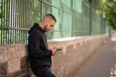 Şehirde sokakta dikilirken yakışıklı bir gencin cep telefonunu kullanırken çekilmiş bir fotoğrafı. Kapüşonlu ve kısa mesajlı kendinden emin bir erkek.. 