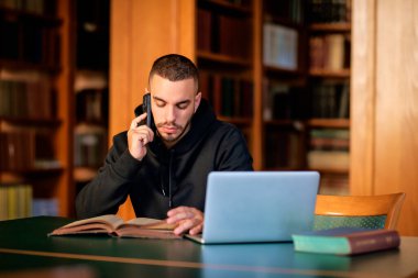Üniversite kütüphanesinde oturan ve öğrenen kendinden emin genç adam. Laptop ve cep telefonu kullanan beyaz bir öğrenci.. 