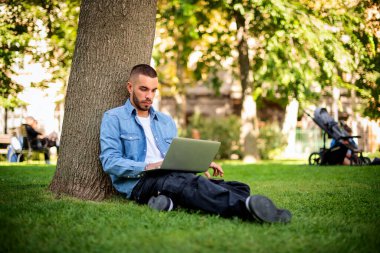 Genç adam dışarıda çalışmak ya da çalışmak için dizüstü bilgisayar kullanıyor. Genç bir erkek günlük kıyafetler giyip parktaki çimlere oturuyor ve internetten öğreniyor. Tam boy çekim.