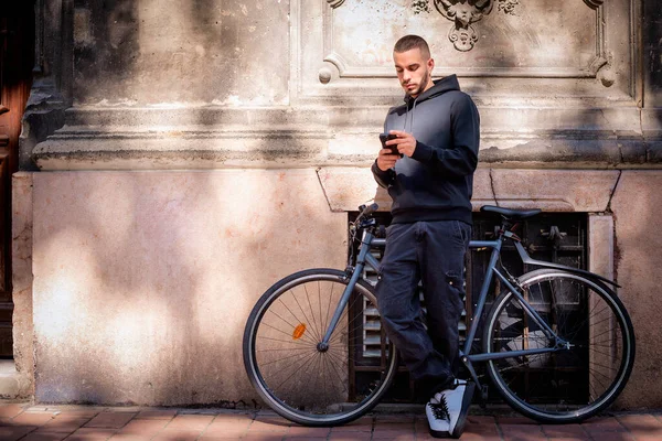 一个英俊的年轻人站在城里的街上用他的手机打了整整一个小时的电话 年轻男子骑着自行车在墙边放松 发短信 — 图库照片