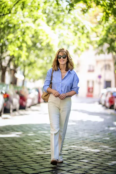 市街地を歩いている美しい中年の女性 白いズボンで青いシャツを着ているブロンドハイレッドの女性 全長について — ストック写真