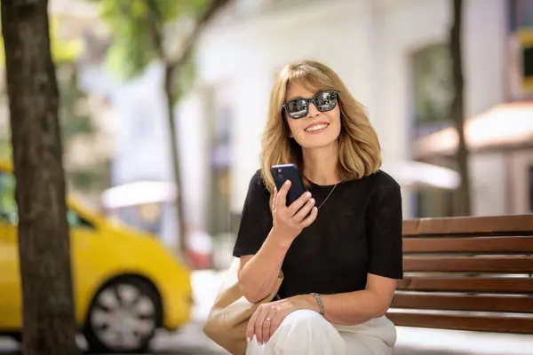迷人的金发女人坐在街上的长椅上 用手机 笑容可亲的女人戴着太阳镜打电话给我 — 图库照片