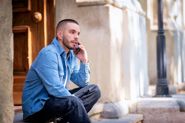 도시의 거리에 앉아있는 자신의 휴대폰을 사용하여 잘생긴 젊은이의 캐주얼 전화하는 스톡 이미지