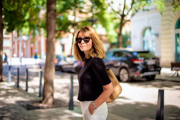 市街地を歩いている美しい中年の女性 白いズボンが付いている黒いシャツを身に着けているブロンド髪の女性 ロイヤリティフリーのストック画像