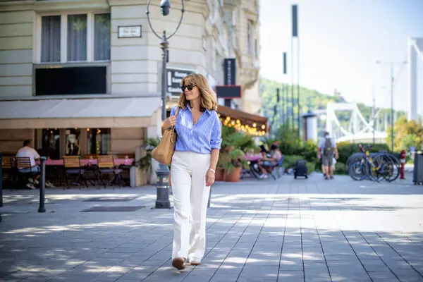 美丽的中年妇女走在城市街道上 金发女郎 身穿蓝色衬衫 图库图片