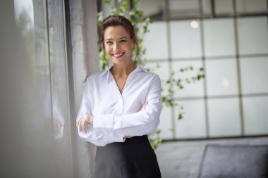 Kahverengi saçlı orta yaşlı bir kadının portresi. Beyaz gömlek ve siyah pantolon giyiyor ve pencerenin yanında duruyor. Çekici bir kadın kameraya bakıyor ve gülümsüyor.. 