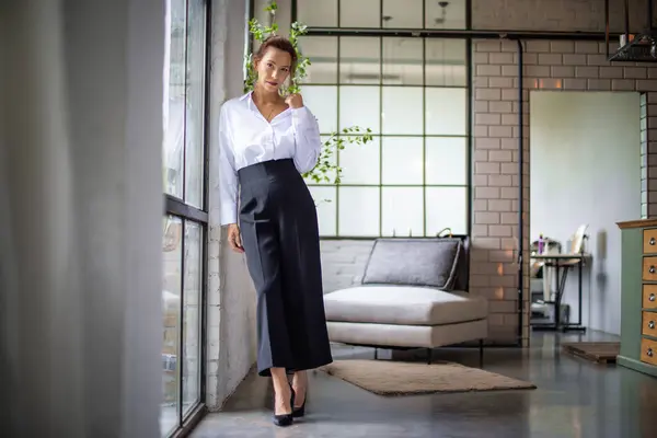 stock image Portrait of a brown haired middle-aged woman wearing a white shirt and black pants and standing by the window. Attractive female looking at camera and smiling. Full length shot. 