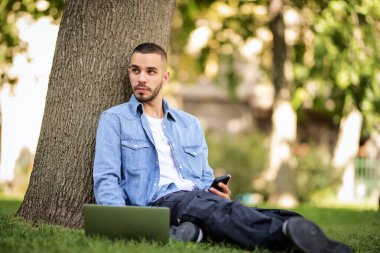 Genç adam dışarıda çalışmak ya da çalışmak için dizüstü bilgisayar kullanıyor. Genç bir erkek günlük giysiler giyip halka açık bir parkta çimlere oturuyor ve mesajlaşıyor. Tam boy çekim.