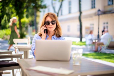 Orta yaşlı sarışın bir kadın bir kafede terasta oturuyor ve dizüstü bilgisayar kullanıyor. İş için dizüstü bilgisayar ve cep telefonu kullanan gülümseyen kadın.. 