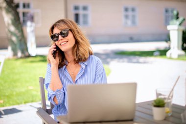 Orta yaşlı sarışın bir kadın bir kafede terasta oturuyor ve dizüstü bilgisayar kullanıyor. Gülümseyen neşeli kadın iş için dizüstü bilgisayar ve cep telefonu kullanıyor..  