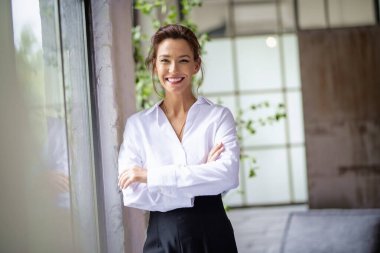 Kahverengi saçlı orta yaşlı bir kadının portresi. Beyaz gömlek ve siyah pantolon giyiyor ve pencerenin yanında duruyor. Çekici bir kadın kameraya bakıyor ve gülümsüyor.. 