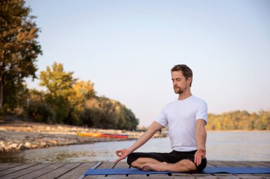 Göl kenarındaki iskelede yoga yapan beyaz bir adam. Olgun bir adam nilüfer pozisyonu ve dinlenme pratiği yapıyor.. 