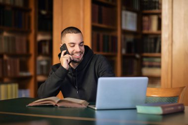 Üniversite kütüphanesinde oturan ve öğrenen kendinden emin genç adam. Laptop ve cep telefonu kullanan beyaz bir öğrenci.. 