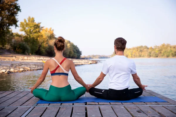一个女人和一个男人坐在瑜伽垫上的荷花姿势的后视镜 在河边的码头上沉思了一长串 — 图库照片