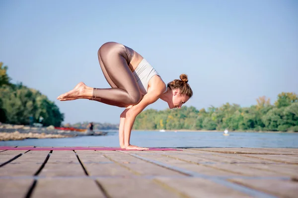 Young woman in crow pose overlooking the river. Full length of a female yoga coach practicing yoga on the pier on riverside.
