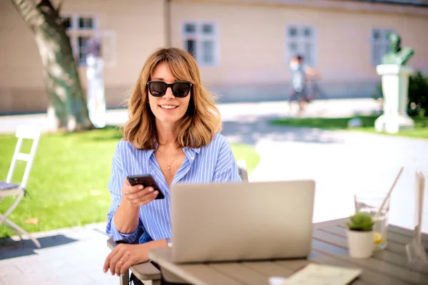 Confident businesswoman sitting at outdoor cafe and working. Attractive female using laptop and text messaging.