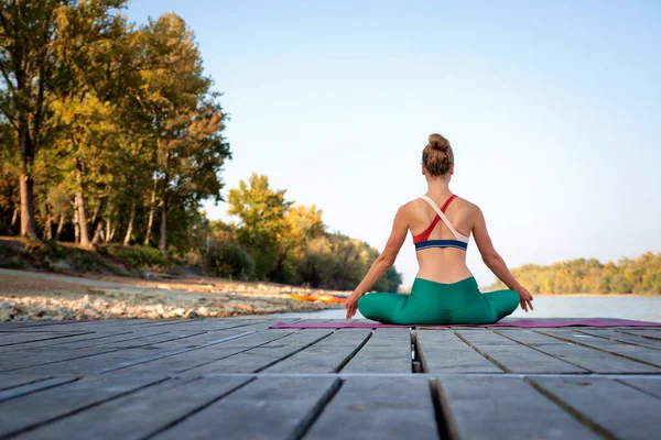 一个女人坐在瑜伽垫子上的荷花姿势的后视镜 全身上下都是一位女性瑜伽教练 在河边的码头上沉思 免版税图库图片