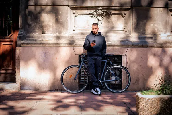 一个英俊的年轻人站在城里的街上用他的手机打了整整一个小时的电话 年轻男子骑着自行车在墙边放松 发短信 免版税图库图片