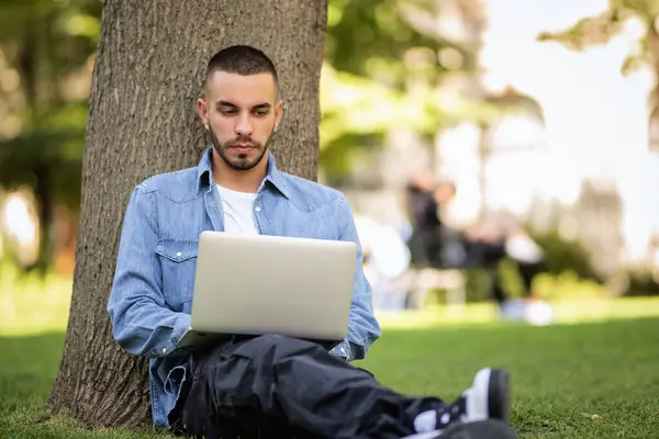 年轻人在室外学习或工作时使用笔记本电脑 年轻男性穿着休闲装 坐在公园的草地上 全长射击 免版税图库照片