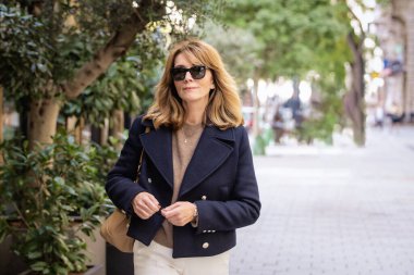 Şehir merkezinde orta yaşlı, sarı saçlı bir kadın caddede yürüyor. Güneş gözlüğü ve mavi ceket giyen çekici bir kadın.. 