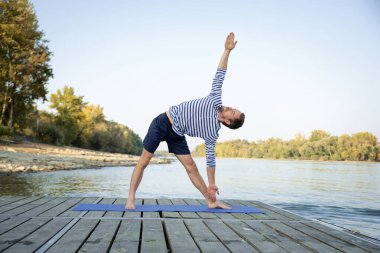 Dışarıda yoga yapan orta yaşlı bir adam. Kafkasyalı adam yoga minderi kullanıyor ve doğayı geriyor..