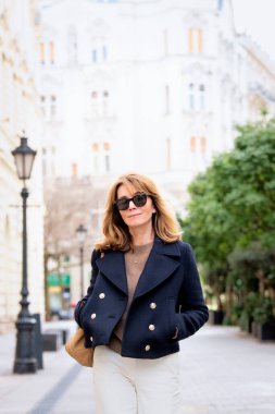 Şehir caddesinde yürüyen orta yaşlı güzel bir kadın. Sarı saçlı, mavi ceketli ve güneş gözlüklü bir kadın.. 