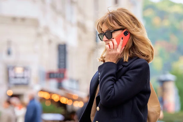 漂亮的中年妇女走在城市街道上 用手机 金发女郎 身穿蓝色外套 戴着太阳镜 — 图库照片