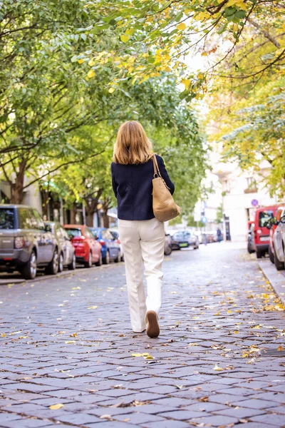 一个中年妇女走在城市街道上的后视镜 金发女人穿着蓝色外套和白色裤子 无法辨认的人 图库图片