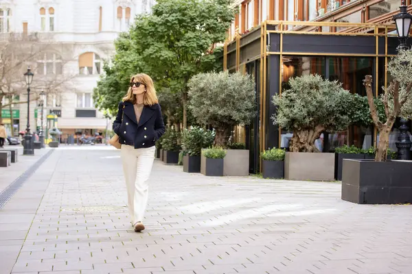 在市中心的街上走着一位金发的中年妇女 迷人的女性 戴着太阳镜 身穿蓝色外套 全长射击 图库图片