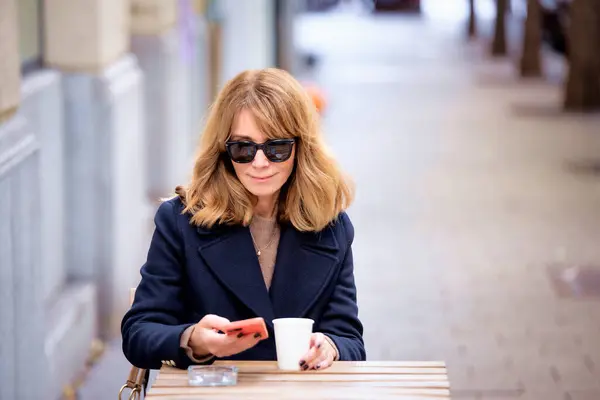一位中年妇女坐在咖啡店外面的桌子旁 一边喝着一杯咖啡 一边用她的智能手机 穿着休闲装和太阳镜的自信女性 免版税图库照片