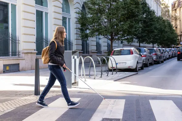 街の道路を横断する白い杖を持つ盲目の女性の肖像画 視覚障害のある女性がカジュアルな服を着て 道を渡るために杖を使った 全長ショット ストックフォト