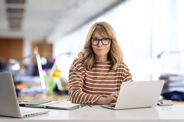 ブロンドハイレッドの実業家がオフィスに座り 仕事のためにラップトップを使用しています メガネとカジュアルな服を着ている自信のあるプロフェッショナル女性 — ストック写真