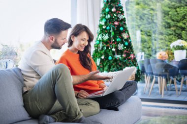Mutlu bir çiftin Noel zamanı koltukta birlikte dinlenmesi. Neşeli kadın ve yakışıklı adam internette gezinirken dizüstü bilgisayar kullanıyor..