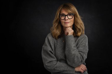 Siyah arka planda olgun bir kadının stüdyo portresi. Sarı saçlı, gri kazaklı ve gözlüklü bir kadın. Kameraya bakıyor ve gülümsüyor. Boşluğu kopyala.