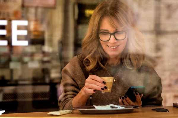 Smiling blond haired woman sitting in cafe and using her smartphone. Attractive female drinking coffee and photographed through the window.