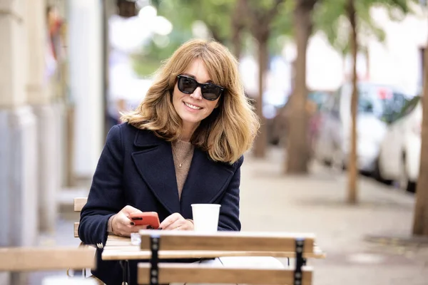 カフェの外のテーブルに座って コーヒーを飲みながらスマートフォンを使用する女性 カジュアルな服やサングラスを着ている自信のある女性 ロイヤリティフリーのストック写真