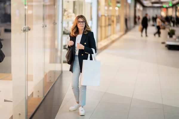 店の窓の前を歩き ショッピングバッグを手に持って ブロンド髪の女性は ブレザーとジーンズを着用 ストック写真