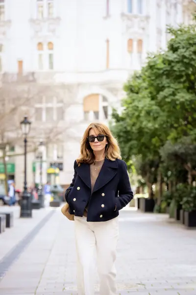 ブロンドの髪をした中年の女性が市内中心部の通りを歩いている 魅力的な女性がサングラスとブルーコートを着用 ストック画像
