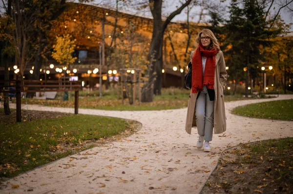 秋にパブリックパークを歩く中年の女性の全長 スカーフとトレンチコートを身に着けているブロンド髪の女性 ストック画像