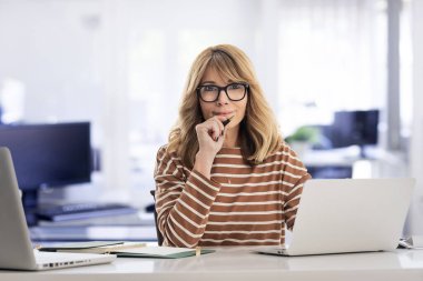 Sarışın, orta yaşlı iş kadını ofiste oturuyor ve iş için dizüstü bilgisayar kullanıyor. Gözlüklü ve günlük kıyafetler giyen kendine güvenen profesyonel bir kadın.. 