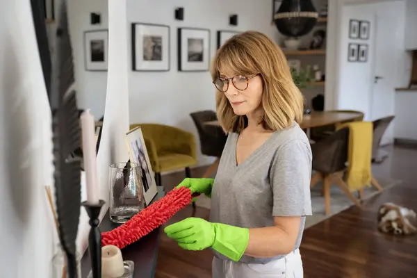 一位中年妇女戴着橡胶手套 手里拿着清洁用品 站在客厅里 微笑的女性清扫家里的壁炉 免版税图库照片
