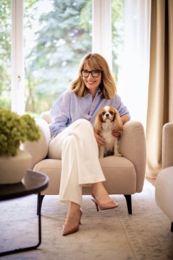Uzun boylu, çekici, sarışın bir kadının sevimli köpeğiyle evdeki koltukta dinlenmesi.. 