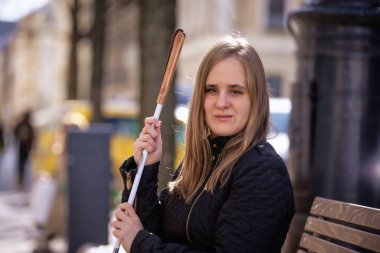 Beyaz baston tutan ve kentteki bir bankta oturan görme engelli bir kadının yakın çekimi. Gülümseyen kadın günlük kıyafetler giyiyor.. 