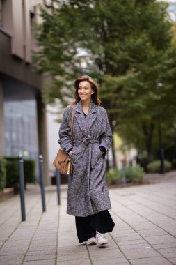 Sonbahar günü şehir caddesinde yürüyen esmer saçlı orta yaşlı bir kadın. Tüvit ceket giyiyor..