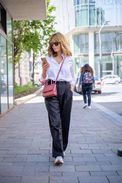 Full length of an attractive woman wearing smart clothes and sunglasses while walking in a city street and text messaging.